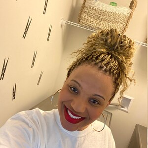 May include: A smiling person with braided hair and large hoop earrings. The person is wearing a white t-shirt and red lipstick. The background features a wall with black clothespin decals and a shelf with a woven basket.