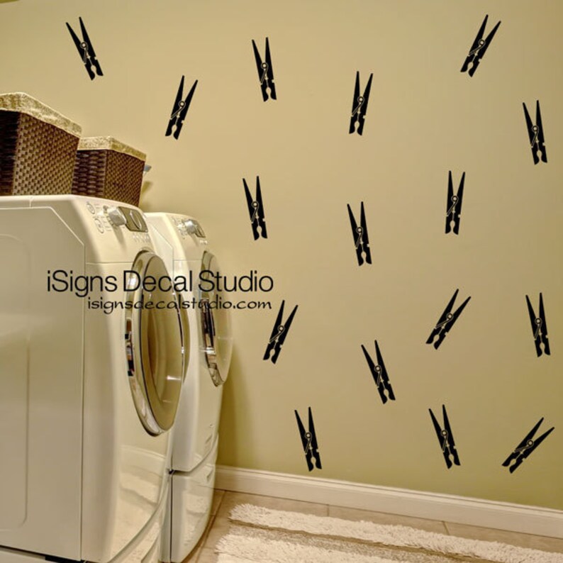 May include: Black clothespin wall decals scattered on a beige wall. The laundry room scene includes a white washing machine and dryer, and two wicker baskets. The text "iSigns Decal Studio" and website address are visible.