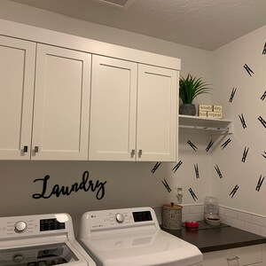 May include: A bright, modern laundry room with white cabinets, a washing machine, and a dryer. The word "Laundry" is displayed in cursive on the wall. Black clothespin decals decorate the wall. A small shelf holds a plant and a sign that says "wash dry fold repeat."