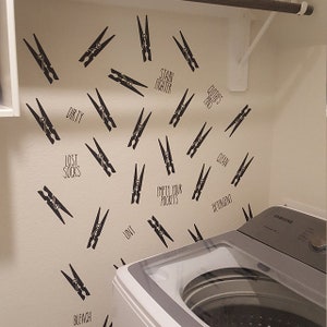 May include: Laundry room wall decor featuring black clothespin decals and text labels like "Dirty," "Clean," and "Lost Socks." The decals are scattered across the wall, creating a playful and functional design element near a washing machine.