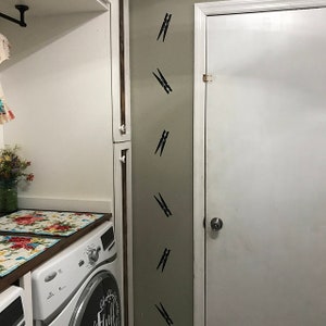 May include: A laundry room interior with a white washing machine and a white door. The wall features several black clothespin decals. Floral placemats sit on the countertop above the washing machine. A jar of flowers is visible.