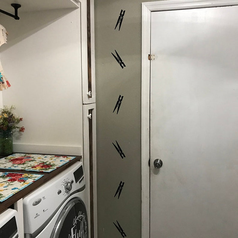 May include: A laundry room interior with a white washing machine and a white door. The wall features several black clothespin decals. Floral placemats sit on the countertop above the washing machine. A jar of flowers is visible.