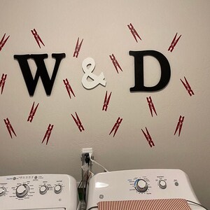 May include: A wall decoration featuring the letters "W & D" in black and white, surrounded by red and white clothespins. The letters are above two white washing machines.