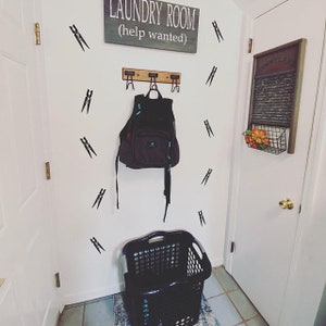 May include: A laundry room scene with a sign that reads "LAUNDRY ROOM (help wanted)". A backpack hangs from a wooden rack. Black clothespin decals decorate the white wall. A black laundry basket sits on the floor.