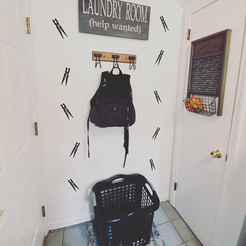 May include: A laundry room scene with a sign that reads "LAUNDRY ROOM (help wanted)". A backpack hangs from a wooden rack. Black clothespin decals decorate the white wall. A black laundry basket sits on the floor.