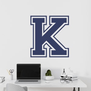 May include: A navy blue letter "K" with a white outline is affixed to a white wall. The letter is in a bold, collegiate style. The image also shows a desk with a laptop, stationery, and decorative items.
