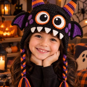 May include: A black crocheted monster hat featuring two orange and purple striped horns, large white and orange eyes, purple bat-like ears, and four white teeth. The hat includes two long braided earflaps with orange, purple, and black tassels.