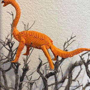 May include: A bright orange dinosaur figurine crafted from tightly woven ribbon, featuring a long neck, a slender tail, and a small black eye. The textured surface of the creature is created by a repeating herringbone pattern of folded fabric.