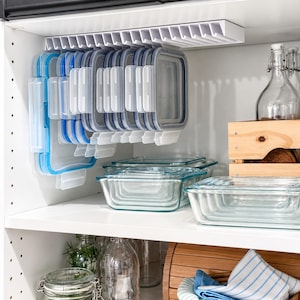 May include: A white plastic under-shelf storage rack holds multiple square and rectangular food storage lids with blue silicone seals. Below the rack, clear glass food storage containers are stacked on a white shelf, with additional glass jars and a wooden crate nearby.