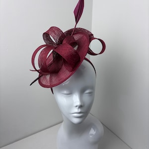 Pode incluir: Um fascinator em tom bordô profundo com uma base circular, detalhes de fitas de sinamay em laços e uma pena vertical. O acessório de cabeça está montado numa tiara fina a condizer, apresentando uma construção em tecido com textura.