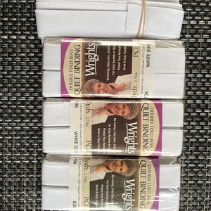 May include: Three packages of white Wrights double fold bias quilt binding, each measuring 3 yards or 2.75 meters long and 0.875 inches wide. The packaging includes text detailing the product name, brand, and material composition of 50% polyester and 50% cotton.