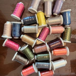 May include: A collection of twenty-five assorted sewing thread spools in various colors including pink, yellow, blue, green, orange, grey, and metallic tones. Each spool features a white plastic base and top with thread wrapped around.