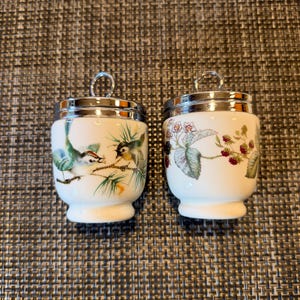 May include: Two white porcelain egg coddlers with silver-tone metal lids and ring handles. One coddler features a colorful illustration of two small birds on a pine branch, while the other displays a botanical print of blackberries and leaves.