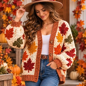 May include: A cream-colored crochet cardigan featuring a button-front closure and ribbed trim in a rust-orange hue. The garment is decorated with various three-dimensional maple leaf appliques in shades of red, orange, yellow, and dark green.