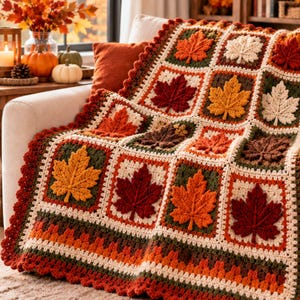 May include: A crochet blanket featuring a grid of square panels, each decorated with a raised, textured maple leaf in shades of red, orange, yellow, brown, and cream. The blanket has a scalloped border in dark red and brown yarn.