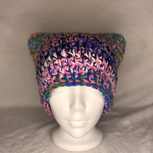 May include: A crochet cat ear beanie featuring a variegated yarn pattern in shades of purple, pink, blue, and green. The hat is constructed with a thick, textured stitch pattern and has two distinct pointed corners at the top, creating a cat-like shape.