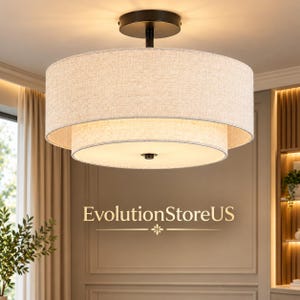 May include: A two-tiered drum ceiling light fixture with a beige fabric shade and a black metal canopy. The light emits a warm glow from within. The text EvolutionStoreUS is printed in gold lettering below the light fixture.