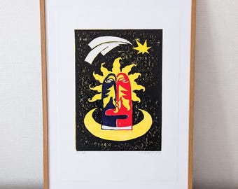 May include: A framed print featuring a stylized sun face with one half dark blue and the other half red, set against a black background with yellow stars, a crescent moon, and a white comet. The artwork is enclosed in a light wood frame.