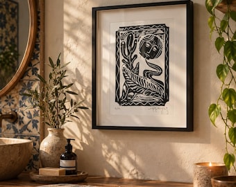 May include: A framed black and white linocut print featuring a stylized figure with a circular head, surrounded by botanical elements like a cactus and fern, enclosed in a decorative border, displayed in a black frame with a white mat.
