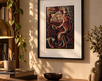 May include: A framed print featuring a stylized, multi-colored woodcut-style illustration of a mythical creature with an open mouth and long tongue, set against a dark background with red and yellow accents, signed and numbered 02/05 at the bottom.