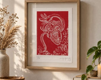 May include: A framed art print featuring a red linocut illustration of a stylized creature with swirling lines and a toothy grin. The print is signed and numbered 1/50 at the bottom, encased in a light-toned wooden frame with a white mat border.