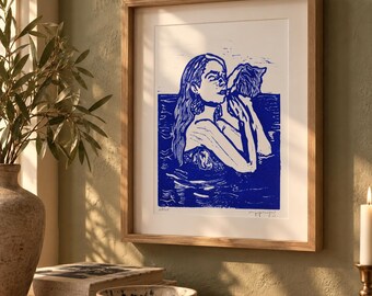 May include: A framed art print featuring a blue and white linocut illustration of a person with long hair holding a cat while submerged in water. The artwork is displayed in a light-colored wooden frame with a white mat, showcasing bold lines.
