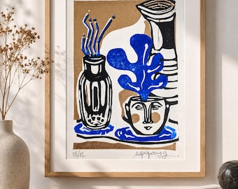 May include: A framed art print featuring a block-print style illustration of two vases and a face-shaped planter with a blue leaf. The artwork uses a palette of black, white, and blue on a tan background, with the handwritten text 08/12 at the bottom.