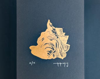 May include: A rectangular print on dark gray paper featuring a gold-colored illustration of a seashell. The print includes the handwritten text 02/02 on the bottom left and a signature on the bottom right, showcasing detailed linework and texture.