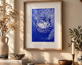 May include: A framed art print featuring a vibrant blue and white abstract linocut design with textured, radiating lines. The artwork is encased in a light-toned wooden frame with a white mat, displayed on a surface with decorative objects.