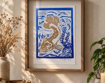 May include: A framed linocut print featuring a tan fish swimming among blue aquatic plants and waves. The artwork is enclosed in a simple wooden frame with a white mat. The print includes a blue border and handwritten text at the bottom.