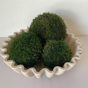 Peut inclure: Un bol peu profond blanc cassé avec un bord festonné et ondulé contenant trois boules de mousse sphériques et texturées d'un vert forêt profond. Le bol présente une finition mate semblable à de la pierre et une large ouverture circulaire.