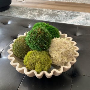 Op de afbeelding: Een ronde, gebroken witte keramische kom met een golvende, geschulpte rand met daarin vijf decoratieve mosballen. De mosballen variëren in textuur en kleur, met tinten van diep bosgroen, helder limoengroen en een bleke, neutrale beige kleur.