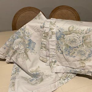 Puede incluir: Una funda de almohada o artículo de ropa de cama de tela blanca con un patrón floral repetitivo en tonos azul suave y verde apagado. El tejido muestra ilustraciones detalladas de rosas en flor y follaje, acabado con un borde sencillo con dobladillo.