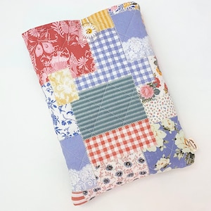 May include: A rectangular quilted fabric pouch featuring a patchwork design of various floral, gingham, and striped patterns in red, blue, yellow, and white. The pouch is secured with a clear button shaped like a white daisy flower.