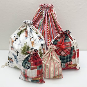 May include: A collection of five fabric drawstring bags in varying sizes. One bag features a snowman and pine tree print, another has vertical stripes, and two feature red and green plaid patterns. One small bag has a vertical floral stripe.