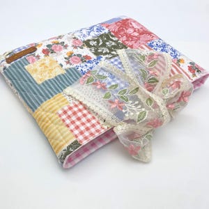 May include: A quilted fabric cover featuring a patchwork design of floral, gingham, and striped patterns in red, blue, yellow, and green. A sheer, cream-colored lace ribbon with embroidered pink flowers and green leaves is tied around the cover.