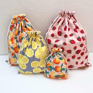 May include: Four drawstring bags of varying sizes featuring fruit patterns. One bag has an orange print, one has a lemon print on a blue and white striped background, one has a strawberry print on a pink background, and one has an orange print.