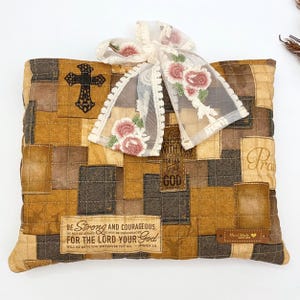 May include: A square quilted pillow featuring a patchwork design in shades of brown and tan. The fabric displays a black cross, deer silhouettes, and the text "Be strong and courageous. Do not be afraid. Do not be discouraged. For the Lord your God will be with you wherever you go. Joshua 1:9." A sheer white ribbon with embroidered pink roses is tied at the top, and a small brown "Hand Made with love" tag is attached to the bottom right corner.