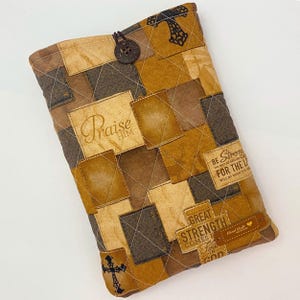 May include: A quilted fabric book cover in shades of brown and tan featuring a patchwork design. It includes a dark brown button closure, black cross motifs, and printed text reading Praise Him, Be Strong, and Great Strength Comes From God.
