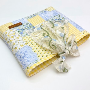 May include: A quilted fabric cover featuring a patchwork design of yellow gingham, blue floral patterns, and white fabric with yellow flowers. A brown rectangular tag is attached to the corner, and a sheer white lace ribbon with green and blue embroidery is tied on top.