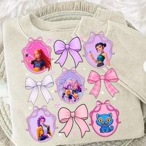May include: A beige crewneck sweatshirt featuring a graphic print of various cartoon characters in oval frames, interspersed with several large pink and white bows. The design includes a blue cat, multiple female figures, and stars.