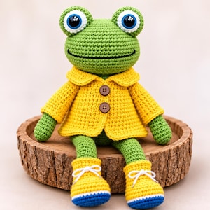 May include: A green crocheted frog plush toy wearing a yellow jacket with two brown buttons and matching yellow boots with white laces and blue soles. The frog has large white eyes with blue irises and black pupils, and a stitched black mouth line.