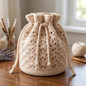 Puede incluir: Un bolso de ganchillo beige con cordón, que presenta un patrón de puntos de concha texturizados en todo el cuerpo. El bolso tiene una abertura superior ajustada por dos cordones trenzados con extremos anudados, creando un borde fruncido.