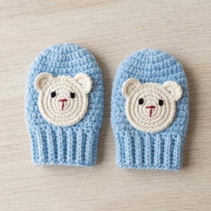 May include: A pair of light blue crochet mittens featuring a cream-coloured bear face appliqué on each. The bear faces have embroidered black eyes and a red stitched nose and mouth. The mittens have a ribbed cuff and a rounded top construction.