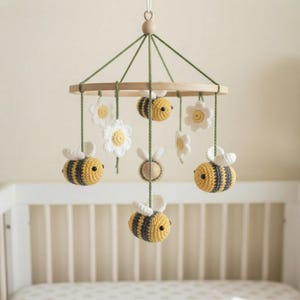 May include: A hanging mobile featuring crocheted yellow and gray striped bees and white flowers with yellow centers suspended from a circular wooden frame by green yarn strings. The structure is held together by a central wooden bead at the top.