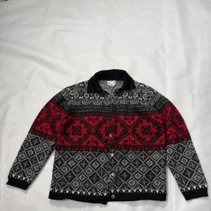 Pode incluir: Um cardigã de manga comprida com botões e um padrão geométrico em preto, vermelho e branco. A peça tem gola, punhos e bainha em preto sólido. A parte frontal apresenta um motivo repetitivo de diamantes e flores com botões prateados.