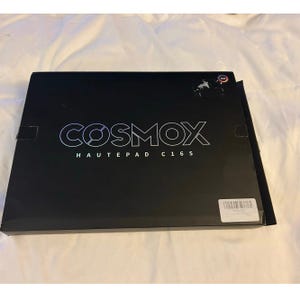 May include: A rectangular black box with the white text COSMOX HAUTEPAD C16S printed on the front. A small circular RGB icon is visible in the top right corner, and a white rectangular barcode sticker is attached to the bottom right corner.