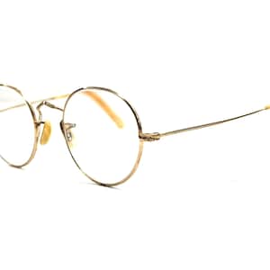 May include: Gold-rimmed eyeglasses with round lenses. The frames are a light gold color, and the temples have a slight curve. The nose pads are a light tan color. The glasses are set against a white background.