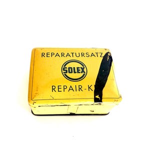 May include: A vintage yellow metal repair kit box. The box features the text "REPARATURSATZ" and "SOLEX REPAIR-KIT" in black. The rectangular box has rounded corners and a black stripe on the side. The box is likely for vehicle repair.