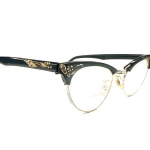 May include: Cat-eye glasses featuring dark blue frames with gold-tone metal lower rims. The upper corners and temples are decorated with embedded clear rhinestones in a floral pattern. The bridge and nose pads are made of gold-tone metal.